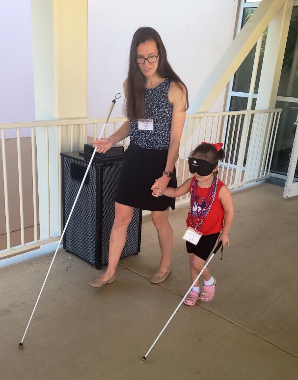 Katy Frets assisting and teaching a young child client, both of them are walking down a hallway using mobility canes
